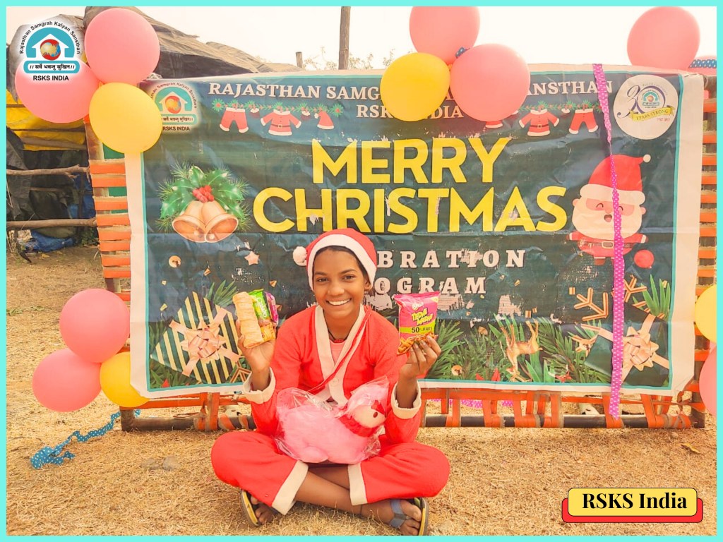 A smiling person dressed in a Santa costume holds snacks and a toy in front of a festive 'Merry Christmas' banner adorned with colorful balloons.