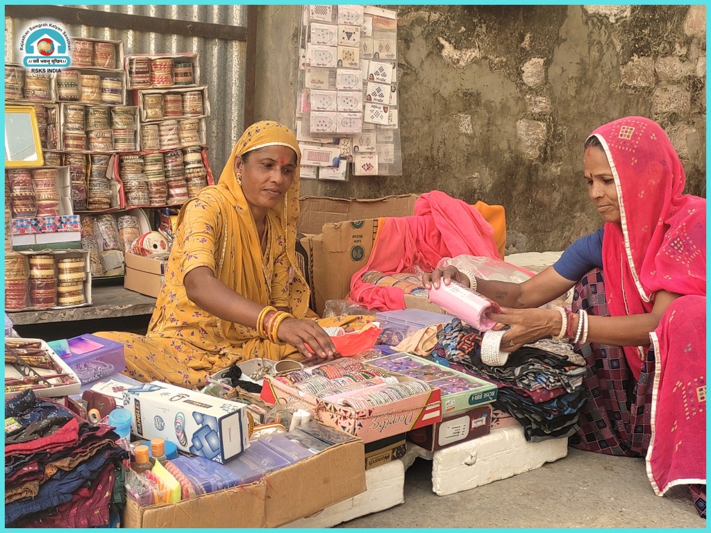 Two women selling colorful fabrics and accessories at a market stall, surrounded by various goods, including bangles and clothing.