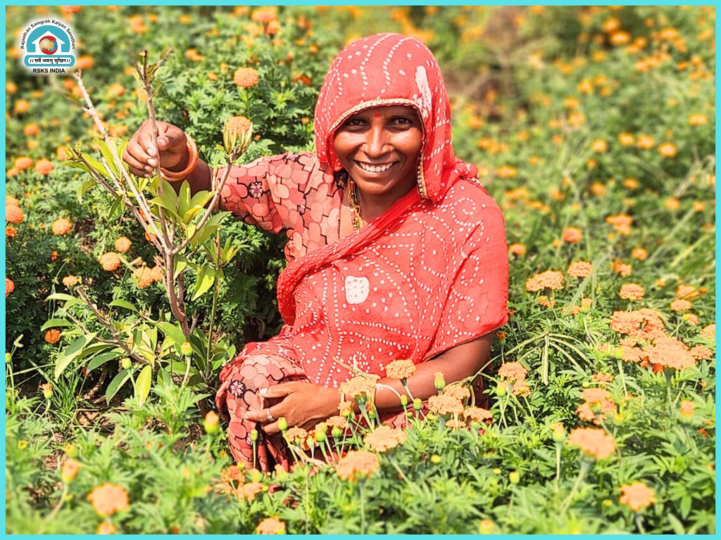 A smiling woman in a red traditional outfit sits among vibrant orange marigold flowers, holding a small plant.