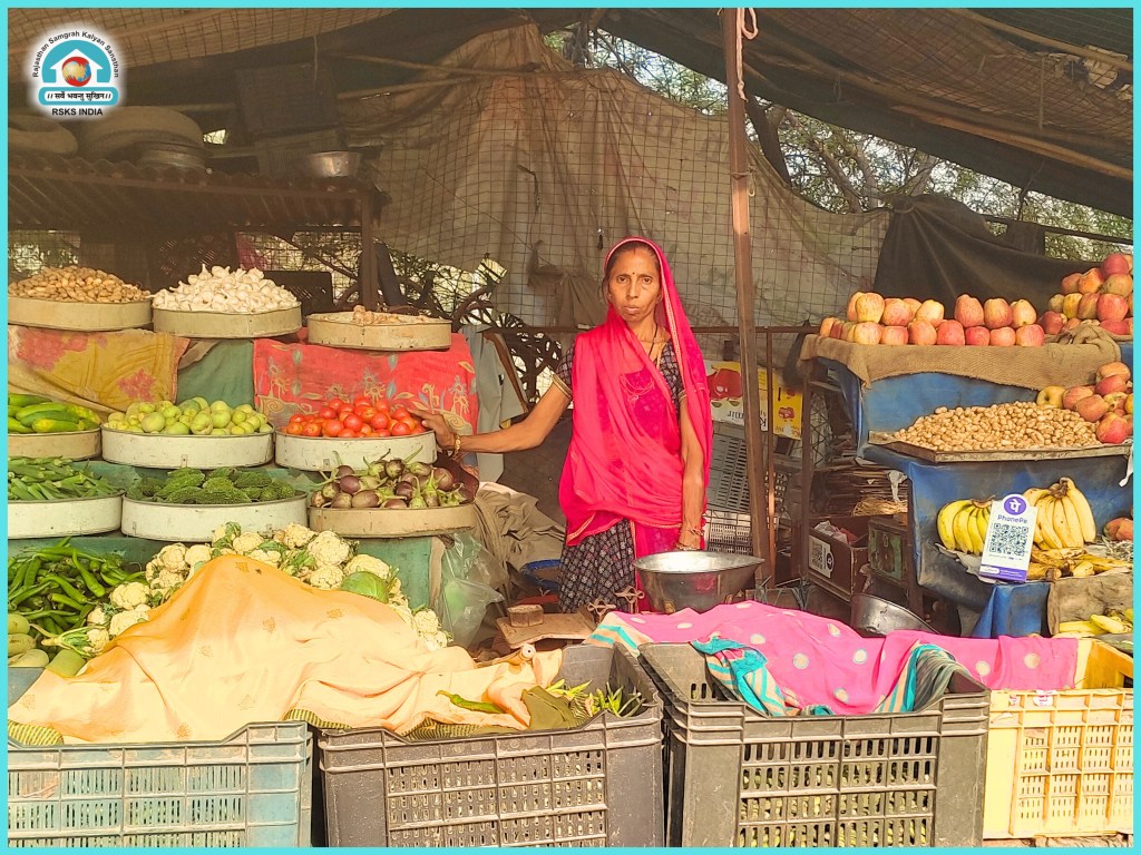 A woman wearing a red scarf stands at a vegetable and fruit market, surrounded by various fresh produce, including green vegetables, fruits, and a basket of goods.