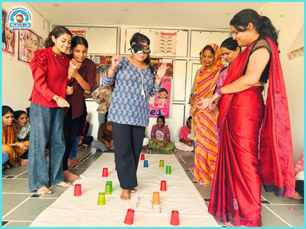 A group of women participating in a fun activity with a blindfolded participant walking over a paper with colored cups, while others cheer and support them in a lively indoor setting.