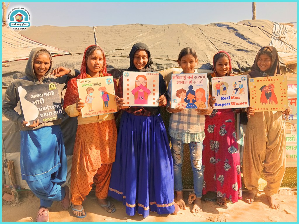 A group of six young women and girls standing together, each holding signs with messages promoting women's rights and empowerment. They are dressed in traditional Indian attire and smiling, with a tent in the background.