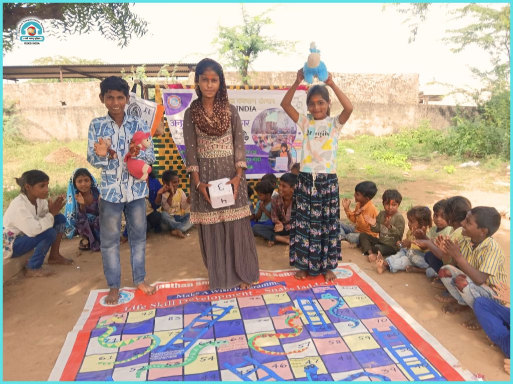 Two children standing on a colorful game mat in an outdoor setting, surrounded by other children sitting on the ground. One child holds a toy and the other raises a plush toy in celebration.