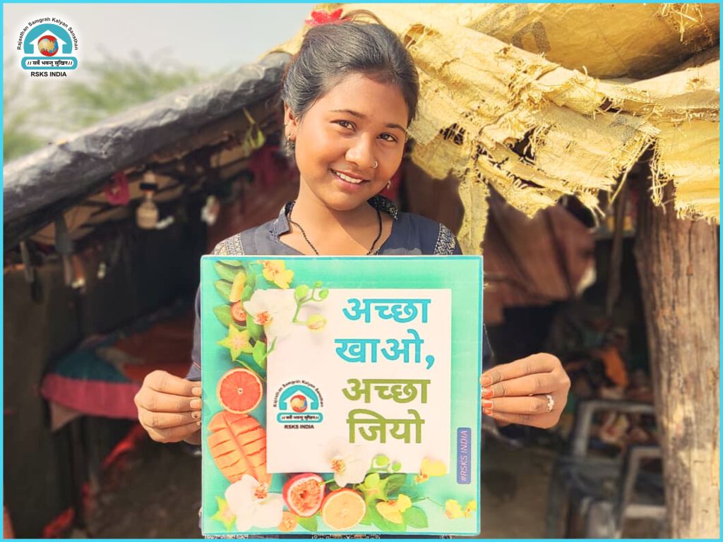 A young girl smiling while holding a sign that reads 'अच्छा खाओ, अच्छा जियो' (Eat well, live well) in a rural setting.