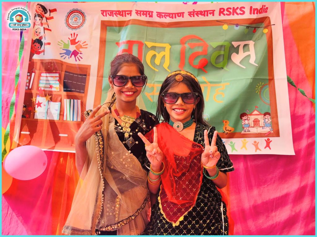 Two smiling girls dressed in traditional attire with sunglasses, posing peace signs in front of a colorful backdrop celebrating a special occasion.