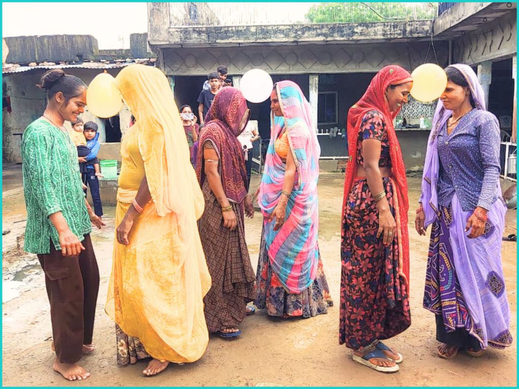 A group of women in colorful traditional clothing are participating in a game involving balloons in an outdoor setting, with children watching in the background.