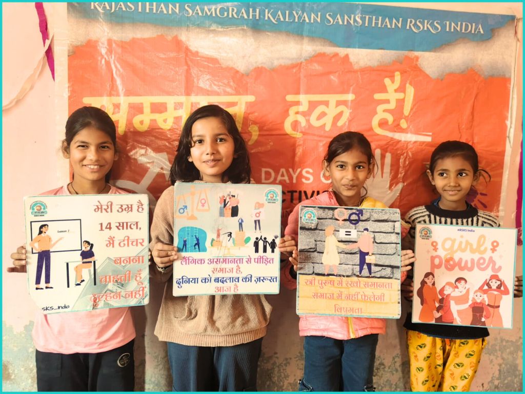 Four girls holding colorful signs with messages about empowerment and education, standing in front of a backdrop that reads 'Rajasthan Samgrah Kalyan Sansthan'.