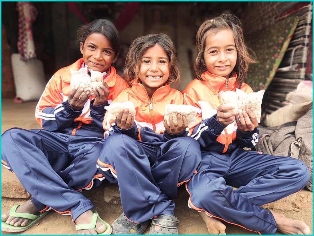 Three smiling children in matching orange and navy tracksuits hold up snacks while sitting on the ground.