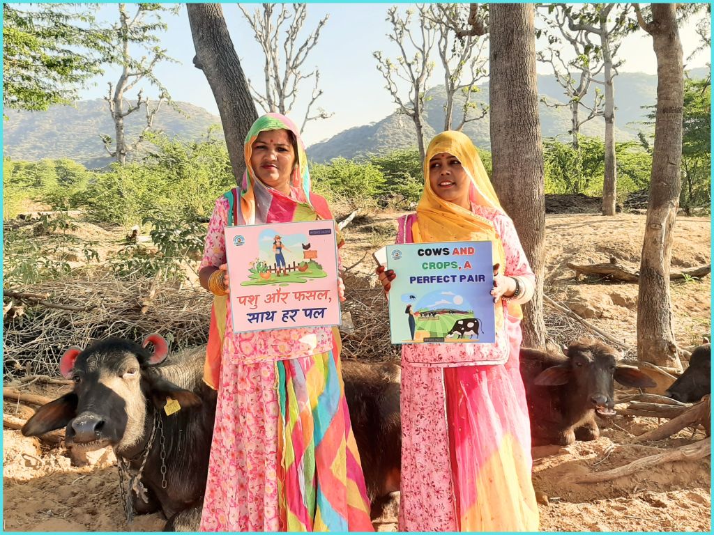 Two women in colorful traditional attire holding signs promoting the connection between cows and crops, with buffaloes in the background and a mountainous landscape.