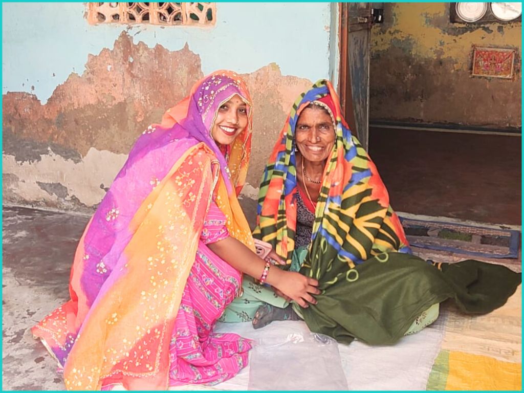 Two women sitting on the floor, smiling and wearing colorful traditional clothing, inside a room with peeling walls.