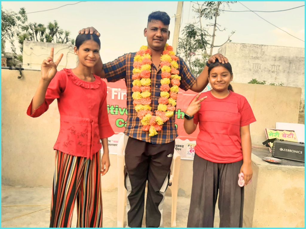 A group of three people celebrates with peace signs. The central figure, wearing a flower garland, stands between two young girls, who are posing happily. The background features a banner and greenery.