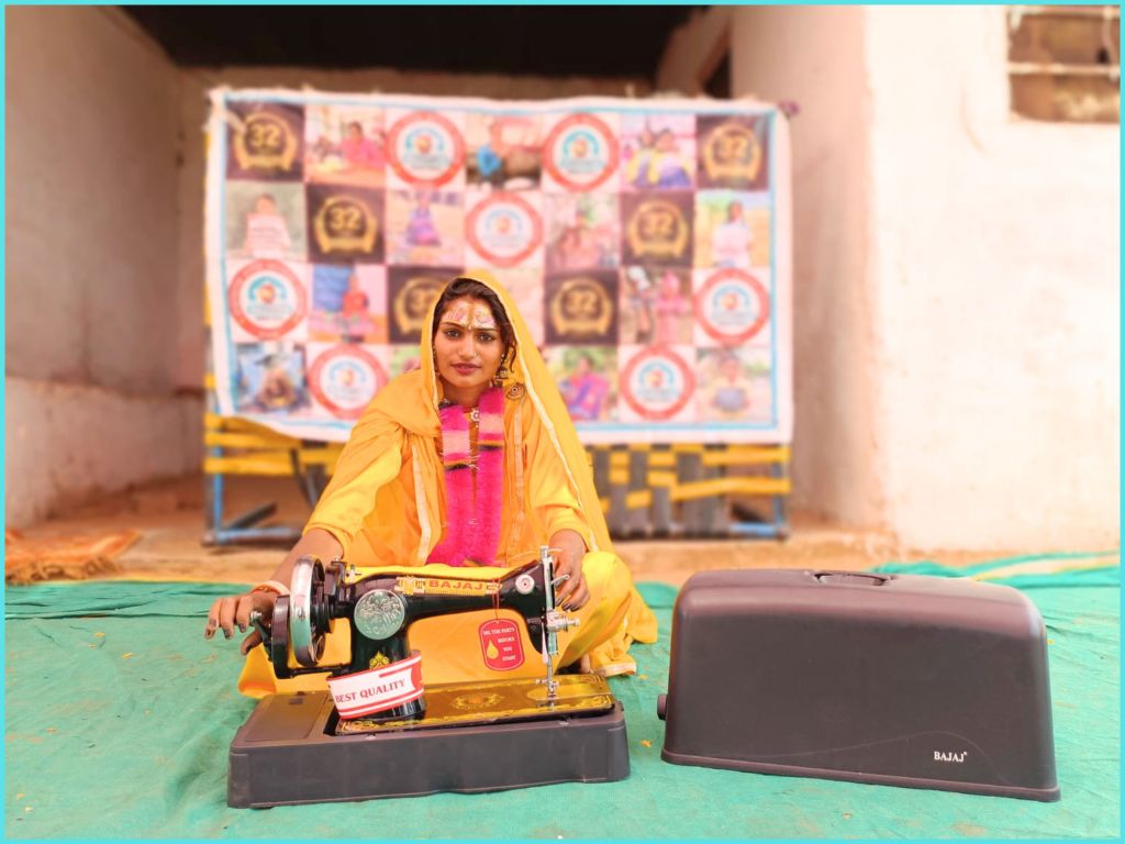 A woman dressed in traditional attire sits beside a sewing machine, smiling. The background features a colorful banner with various images and symbols, on a green floor.