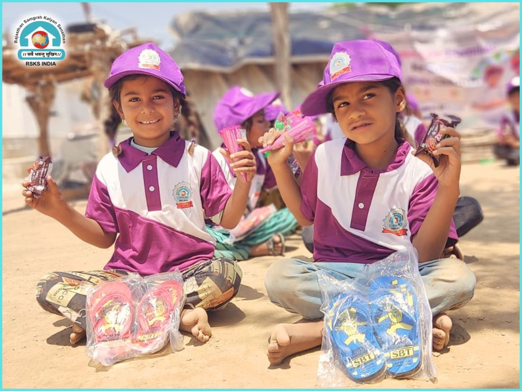 Two young girls sitting on the ground, each holding candy and wearing purple caps. They are dressed in matching purple and white polo shirts and have displayed plastic packages containing colorful flip-flops.