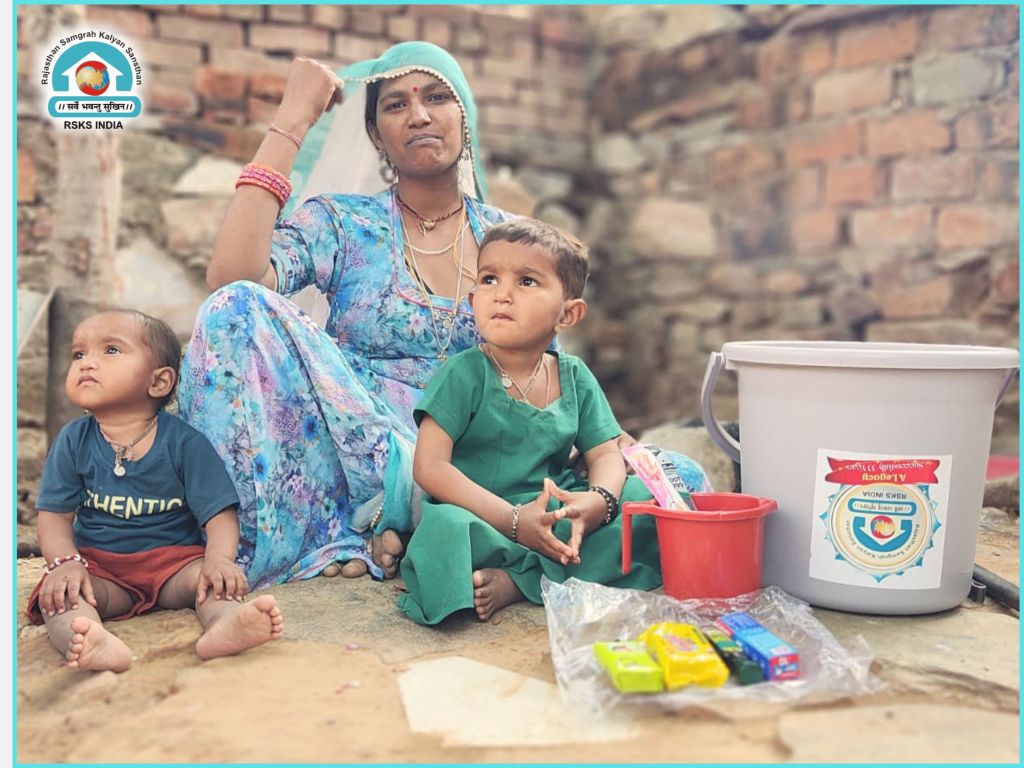 A woman sitting on the ground with two children nearby, one in green clothing and the other in a navy blue shirt, next to a bucket and colorful items on the floor, set against a rustic background.
