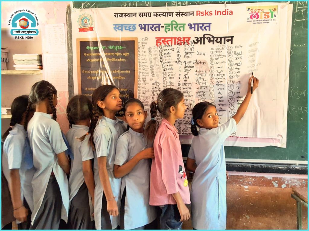 Group of schoolgirls participating in a cleanliness initiative, standing in front of a poster detailing a hygiene campaign. They are engaged in writing on a board with lists related to the campaign.