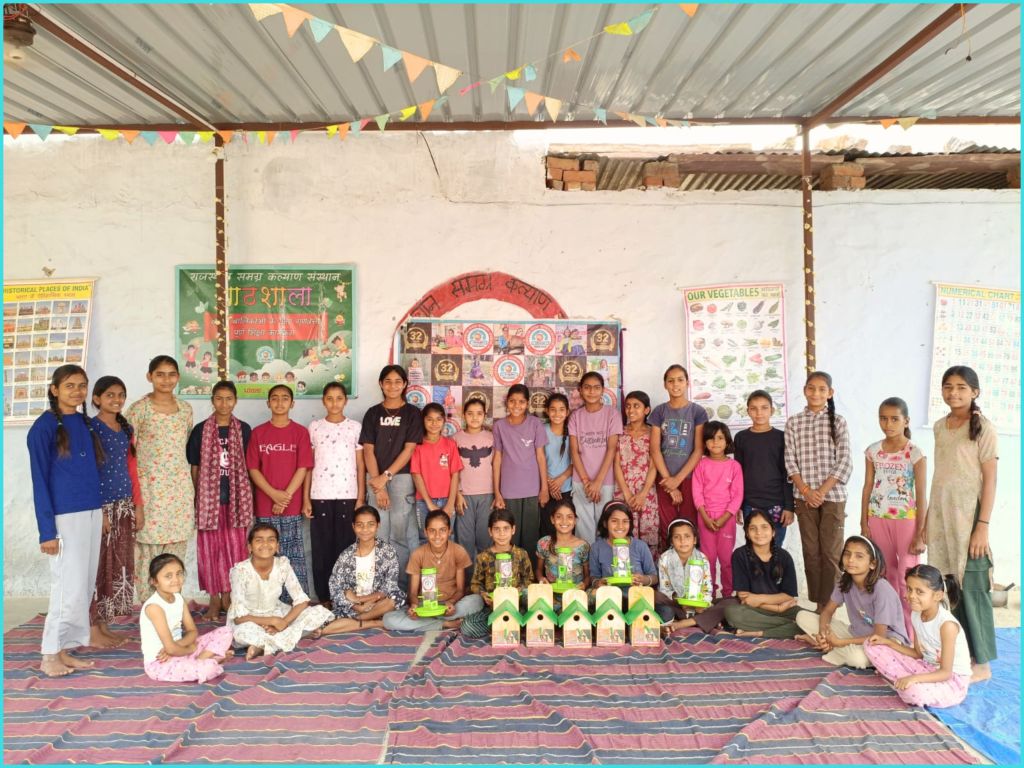 A group of children and young adults posing together in a classroom setting, with colorful educational posters on the walls. Some children are seated on the floor, while others are standing. They appear to be engaged in a school or community project, with green birdhouses displayed in front of them.