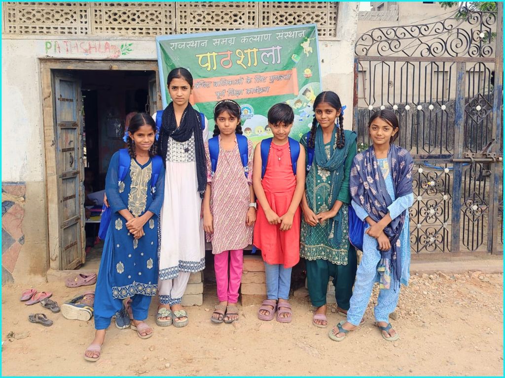 A group of six young girls standing in front of a colorful educational center, wearing traditional attire and backpacks, with a sign reading 'Pathshala' above the entrance.