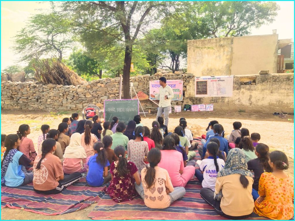 A group of children sitting on the ground in a rural setting, listening to a male instructor teaching in front of a chalkboard. The background features stone walls and trees.