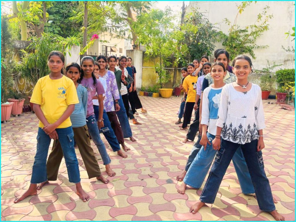 A group of young girls standing in a line outdoors, smiling and dressed in casual clothing, on a patterned floor surrounded by greenery.