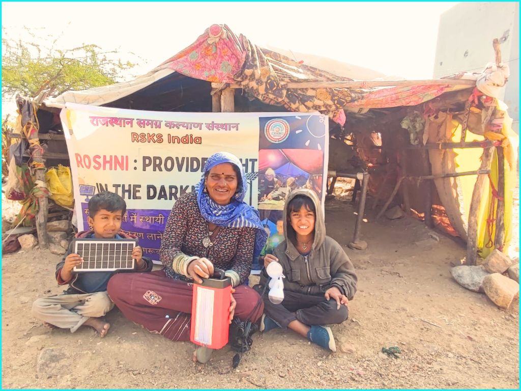 A smiling woman and two children pose in front of a makeshift shelter, with a banner promoting the Roshni project by RSKS India. The woman holds a lantern, while one child displays a solar panel and the other holds a toy.