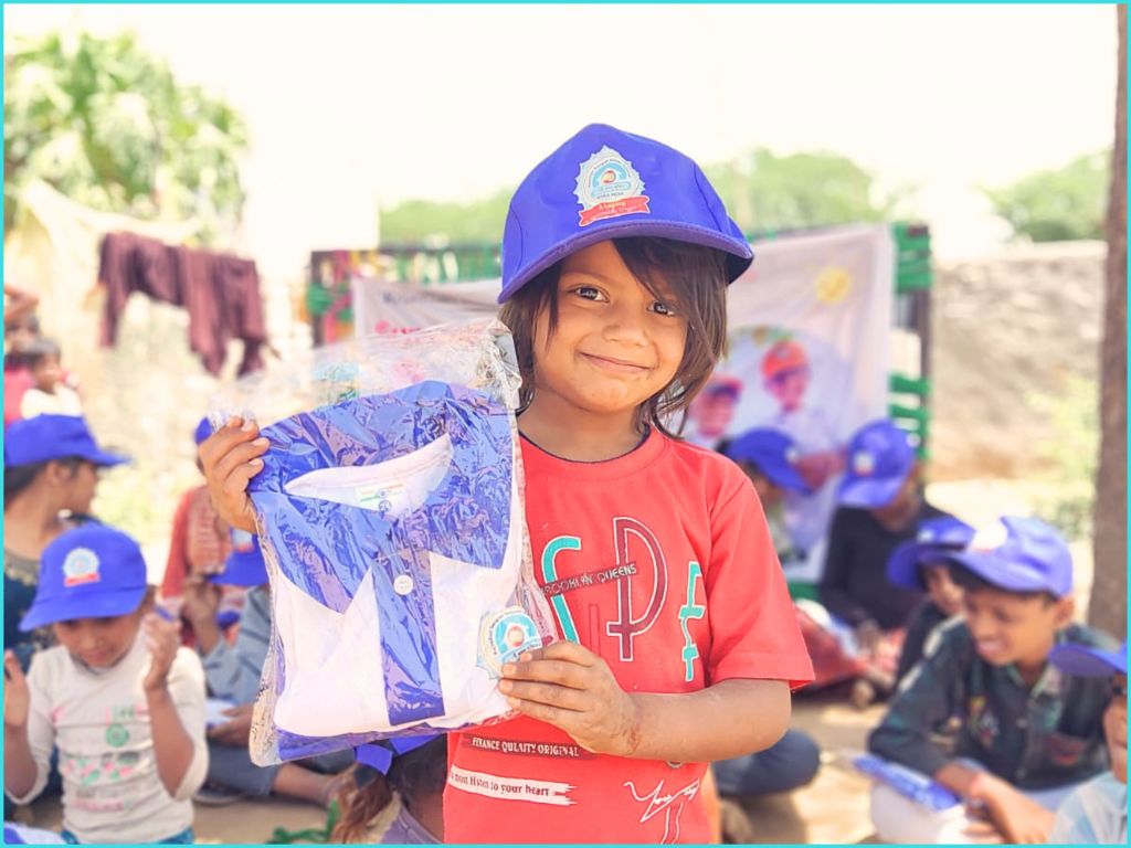 A smiling child holding a new school uniform wrapped in plastic, wearing a red shirt and a blue cap, with other children in the background.