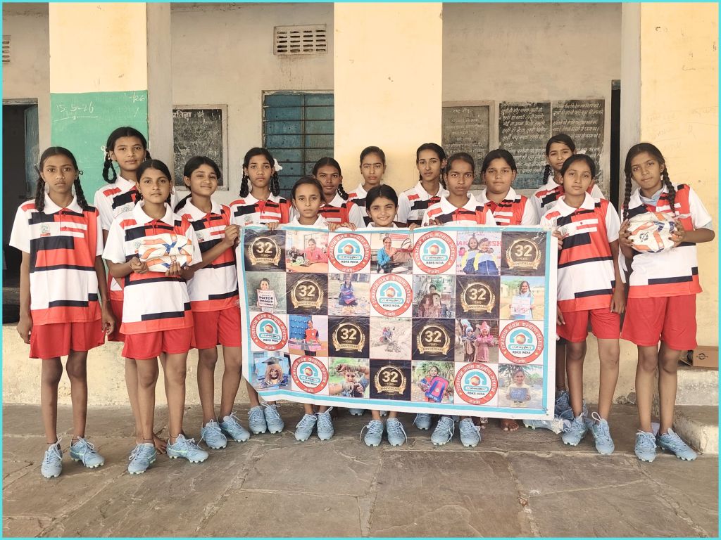 A group of young female athletes posing together in sports uniforms, standing in front of a wall. They are holding a banner featuring various images and logos related to their team or event.