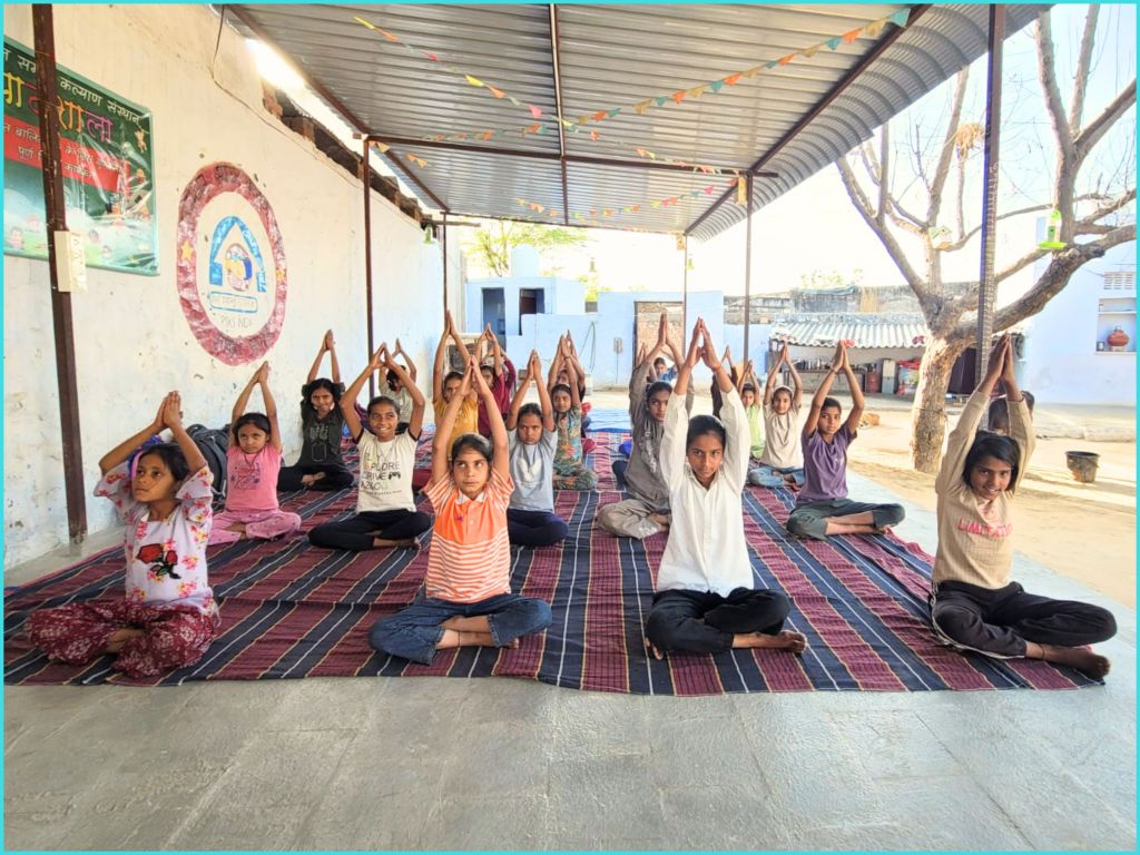 A group of children practicing yoga indoors, seated on a rug with their arms raised overhead in a classroom-like setting.