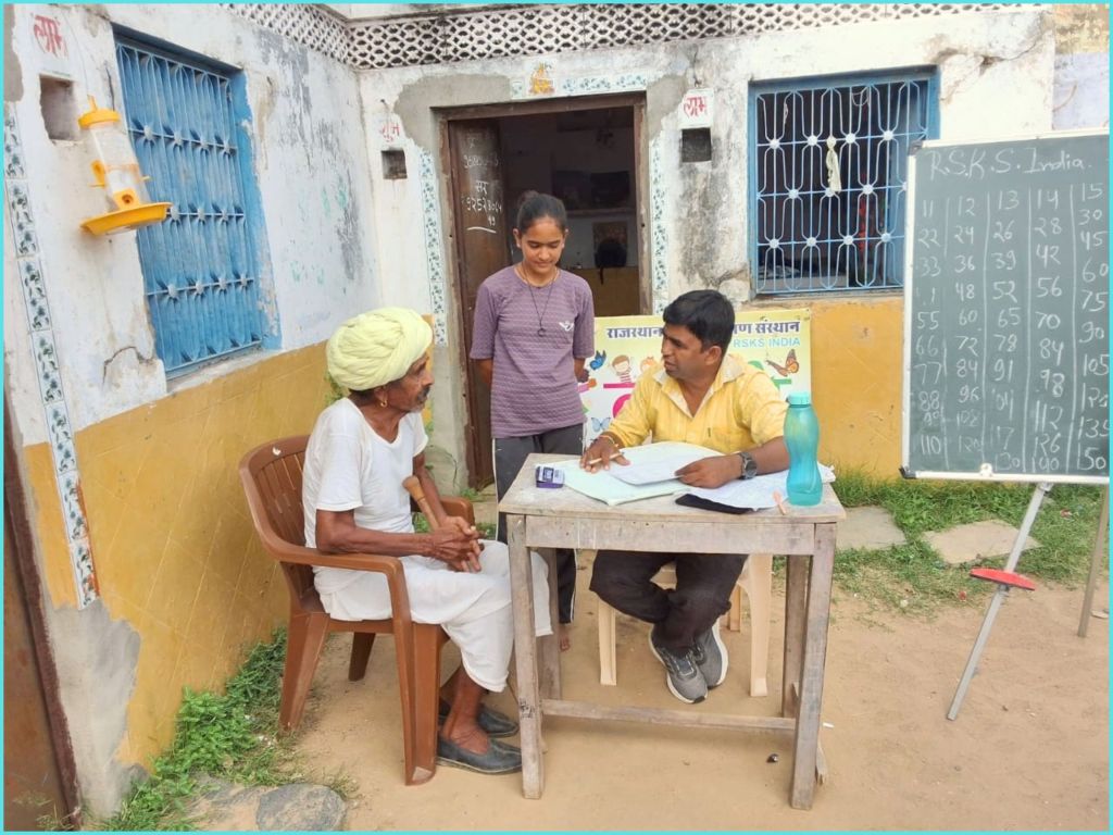 A rural scene showing two men and a woman at a table outside a building. One man, wearing a white shirt and a yellow turban, is seated in a chair, while another man in a yellow shirt is writing notes at the table. The woman stands beside them, observing. A blackboard with numbers is visible in the background, along with colorful signage.